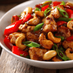 Close-up of authentic cashew chicken stir-fry with diced tender chicken, golden cashews, red bell peppers, sliced green onions, and glossy soy-based sauce presented in a shallow white bowl on a rustic wooden surface