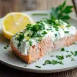 Close-up of a baked salmon fillet topped with creamy white Boursin cheese spread, sprinkled with fresh green parsley and garnished with a lemon wedge on a white plate, highlighting the salmon's pink-orange color and light brown baked edges.