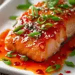 Close-up of a tender baked salmon fillet glazed with sticky sweet chili garlic sauce, garnished with chopped green onions and white sesame seeds on a white rectangular plate