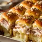 Close-up of baked turkey and cheese sliders in a rustic baking dish showing melted Swiss cheese, golden brown buns, pale pink turkey breast, and a butter Dijon mustard glaze with poppy seeds