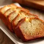 Close-up of classic banana bread loaf sliced into thick pieces on a white ceramic plate, showing moist crumb and golden brown crust with visible banana specks in warm rustic lighting