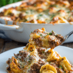 Close-up of a rustic beefy tortellini bake in a white ceramic dish with melted golden mozzarella and parmesan cheese, tender pasta, browned ground beef, tomato marinara sauce, and sprinkled Italian herbs, served on a white plate on a wooden surface.