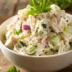Close-up of creamy classic chicken salad with shredded chicken, finely chopped celery and red onion in a shallow white bowl topped with fresh parsley, photographed with warm natural light on rustic surface