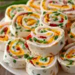 Close-up of cream cheese roll-ups sliced into pinwheels arranged on a white round plate, showing spiral layers of cream cheese, chopped chives, diced red bell pepper, shredded cheddar cheese, and sliced smoked turkey on rustic wooden surface