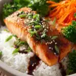 Close-up of pan-seared salmon fillet with crispy golden skin atop fluffy white jasmine rice, garnished with steamed broccoli, carrot ribbons, sesame seeds, and chopped green onions, drizzled with soy-ginger glaze on a rustic surface.