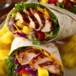 Close-up of Hawaiian BBQ Chicken Wraps cut diagonally showing layers of grilled chicken, cheddar cheese, pineapple chunks, lettuce, red onion, and bell pepper on a white rectangular plate with extra pineapple chunks.