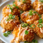 Close-up of crispy bang bang salmon bites with golden panko crust and orange-pink salmon, drizzled with creamy spicy mayo and sweet chili sauce, garnished with sliced green onions and sesame seeds on a white plate