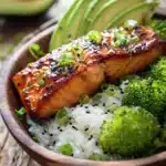 Close-up of a honey glazed pan-seared salmon fillet on a bed of jasmine rice, with steamed broccoli florets and sliced avocado, garnished with sesame seeds and chopped green onions, presented in a rustic bowl under warm natural light.
