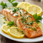 Close-up of tender lemon garlic baked salmon fillet on a white round plate, garnished with lemon slices and fresh parsley, warm natural light highlighting the glossy pink-orange salmon.