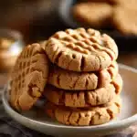 Close-up of stacked peanut butter cookies with crisscross fork marks and visible peanut bits on a white ceramic plate with warm natural lighting