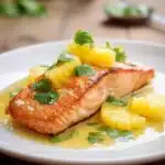 Pan-seared and baked salmon fillet with golden pineapple glaze, garnished with thin pineapple slices and fresh chopped cilantro on a white round plate, showcasing a crisp exterior and moist flaky texture in warm natural light.