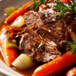 Close-up of sliced tender pot roast with rich brown gravy, carrots, celery, and fresh herbs on an off-white plate with rustic wooden background
