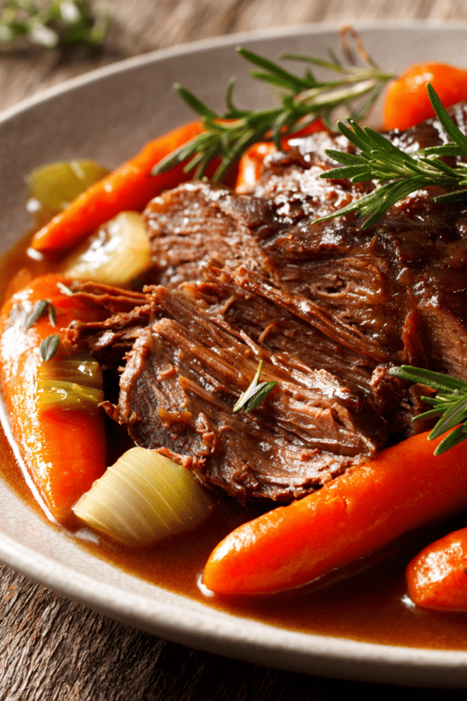 Close-up of sliced tender pot roast with rich brown gravy, carrots, celery, and fresh herbs on an off-white plate with rustic wooden background