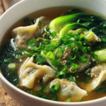Close-up of potsticker soup in a white bowl with tender dumplings, clear golden broth, bright green baby bok choy, sliced green onions, and sesame oil drizzle on rustic surface