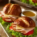 Close-up of two pretzel sandwiches on a white rectangular plate, showing soft pretzel rolls with sliced turkey breast, Swiss cheese, lettuce, and tomato, with a small ramekin of honey mustard sauce on a rustic wooden surface.