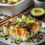 Close-up of spicy salmon sushi bake with golden crispy spicy mayo topping, salmon chunks, avocado slices, green onions, and sesame seeds served on white ceramic plate and rustic wooden surface