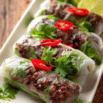 Close-up of crispy pan-fried Thai Basil Beef Rolls wrapped in translucent rice paper with fresh green basil leaves and red chili peppers, served on a white rectangular plate with lime wedges and cilantro garnish.