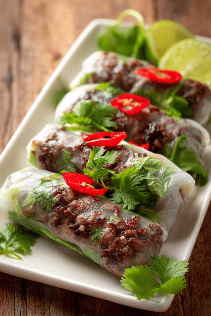 Close-up of crispy pan-fried Thai Basil Beef Rolls wrapped in translucent rice paper with fresh green basil leaves and red chili peppers, served on a white rectangular plate with lime wedges and cilantro garnish.