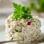 Close-up of a creamy classic tuna salad with finely chopped celery and red onion, neatly scooped on a white plate with a parsley sprig on top, showcasing off-white tuna and pale yellow mayonnaise in warm natural light.