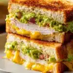 Close-up of a turkey guacamole grilled cheese sandwich cut diagonally, showing crispy toasted sourdough bread, bright green guacamole, melted cheddar cheese, and tender sliced turkey on a white plate