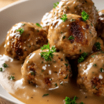 Close-up of tender ground turkey meatballs with visible herbs, covered in smooth thick brown gravy, served on a shallow white round plate with warm rustic background.