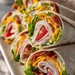 Close-up of turkey ranch club wraps cut diagonally and stacked on a white plate, showing layers of sliced turkey, crispy bacon, lettuce, tomatoes, cheddar cheese, and ranch dressing inside soft flour tortillas.