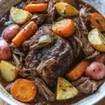 3 packet pot roast slow cooker - the image is a close-up of a bowl of beef stew. the stew is made with chunks of beef, carrots, potatoes, and herbs. the beef is cooked medium-rare and appears to be tender and juicy. the potatoes are cut into small cubes and are arranged in a circular pattern around the beef. the carrots are bright orange and the potatoes are yellow. the herbs are scattered throughout the stew, adding a pop of green color to the dish. the bowl is white with a blue rim and is sitting on a wooden table.