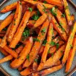 Air fryer garlic sweet potato fries - the image shows a plate of cooked sweet potato fries. the fries are orange in color and have a crispy texture. they are garnished with chopped parsley and sesame seeds. the plate is round and has a dark blue rim. the background is a dark grey countertop.