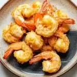 Air fryer shrimp cornstarch - the image shows a plate of cooked shrimp on a white marble countertop. the shrimp are golden brown and appear to be seasoned with herbs and spices. they are arranged in a circular pattern on the plate, with some overlapping each other. the plate is round and has a dark brown rim. the background is blurred, making the shrimp the focal point of the image.