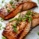 Air fryer teriyaki salmon - the image shows two pieces of salmon on a white plate. the salmon is cooked to a golden brown color and is covered in sesame seeds and green onions. the plate is garnished with chopped green onions, adding a pop of color to the dish. the background is blurred, making the salmon the focal point of the image.