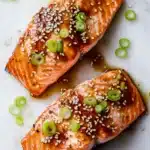 Air fryer teriyaki salmon - the image shows two pieces of salmon on a white marble surface. the salmon is cooked to a golden brown color and is covered in sesame seeds and green onions. the onions are scattered around the salmon, adding a pop of color to the dish. the background is blurred, making the salmon the focal point of the image.
