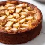 Apple cake one bowl - the image is of a freshly baked apple tart on a white marble countertop. the tart has a golden brown crust and is topped with sliced apples arranged in a criss-cross pattern. the apples are cut into small cubes and appear to be freshly baked. in the background, there is a silver plate with a fork and knife on it. the overall aesthetic of the image is elegant and sophisticated.