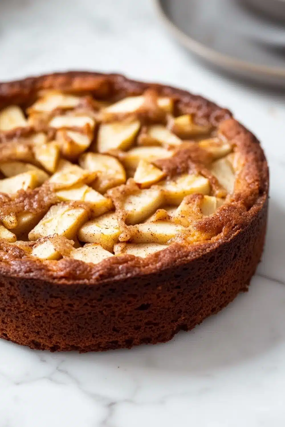 Apple cake one bowl - the image is of a freshly baked apple tart on a white marble countertop. the tart has a golden brown crust and is topped with sliced apples arranged in a criss-cross pattern. the apples are cut into small cubes and appear to be freshly baked. in the background, there is a silver plate with a fork and knife on it. the overall aesthetic of the image is elegant and sophisticated.