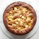 Apple cake one bowl - the image is of a round cake on a white marble countertop. the cake appears to be freshly baked and has a golden brown crust on top. it is topped with sliced apples arranged in a criss-cross pattern, with some of the apples overlapping each other. the apples are cut into small cubes and are arranged in an even layer on top of the cake. the plate it is on is round and white, and the cake is placed on is placed in the center of the image.