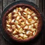 Apple cake yogurt - the image is a close-up of a round cake on a wooden table. the cake appears to be freshly baked and has a golden brown crust on top. the top of the cake is covered in a lattice-like pattern, which is made up of small cubes of apples arranged in a criss-cross design. the apples are evenly spaced and appear to be caramelized. the table has a rustic, weathered appearance, with visible grain and knots. the overall mood of the image is warm and inviting.