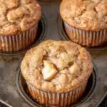 Apple muffins for kids - the image shows a muffin tin filled with freshly baked muffins. the muffins are golden brown in color and have a crumbly texture on top. one of the muffins has a bite taken out of it, revealing a small piece of apple inside. the tin is sitting on a wooden table.