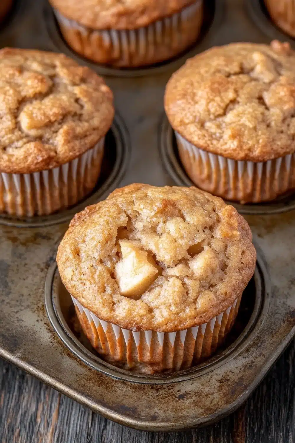 Apple muffins for kids - the image shows a muffin tin filled with freshly baked muffins. the muffins are golden brown in color and have a crumbly texture on top. one of the muffins has a bite taken out of it, revealing a small piece of apple inside. the tin is sitting on a wooden table.