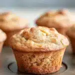 Apple muffins for kids - the image shows a close-up of a freshly baked muffin in a muffin tin. the muffin is golden brown in color and has a crumbly texture on top. it appears to be freshly baked and is sitting on a baking tray. in the background, there are more muffins visible, but they are slightly blurred. the tray is placed on a wooden surface.