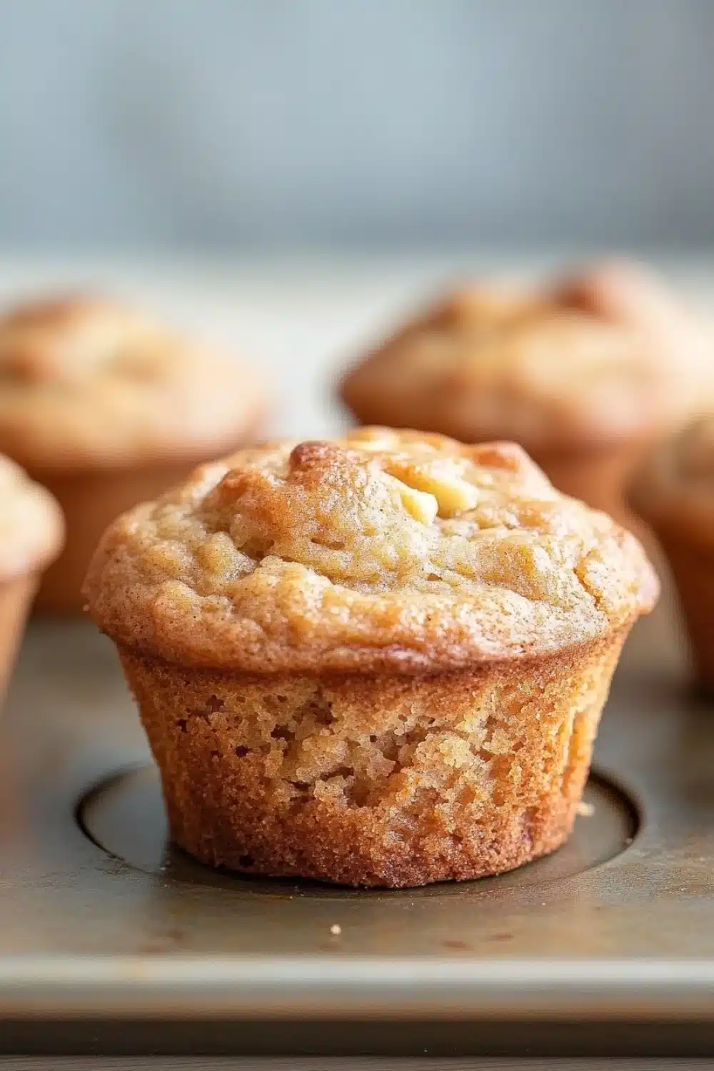 Apple muffins for kids - the image shows a close-up of a freshly baked muffin in a muffin tin. the muffin is golden brown in color and has a crumbly texture on top. it appears to be freshly baked and is sitting on a baking tray. in the background, there are more muffins visible, but they are slightly blurred. the tray is placed on a wooden surface.