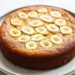 Banana cake - the image is of a round cake on a white plate. the cake appears to be freshly baked and has a golden brown crust. on top of the cake, there are sliced bananas arranged in a circular pattern. the bananas are evenly spaced and appear to be ripe and juicy. the plate is sitting on a marble countertop.