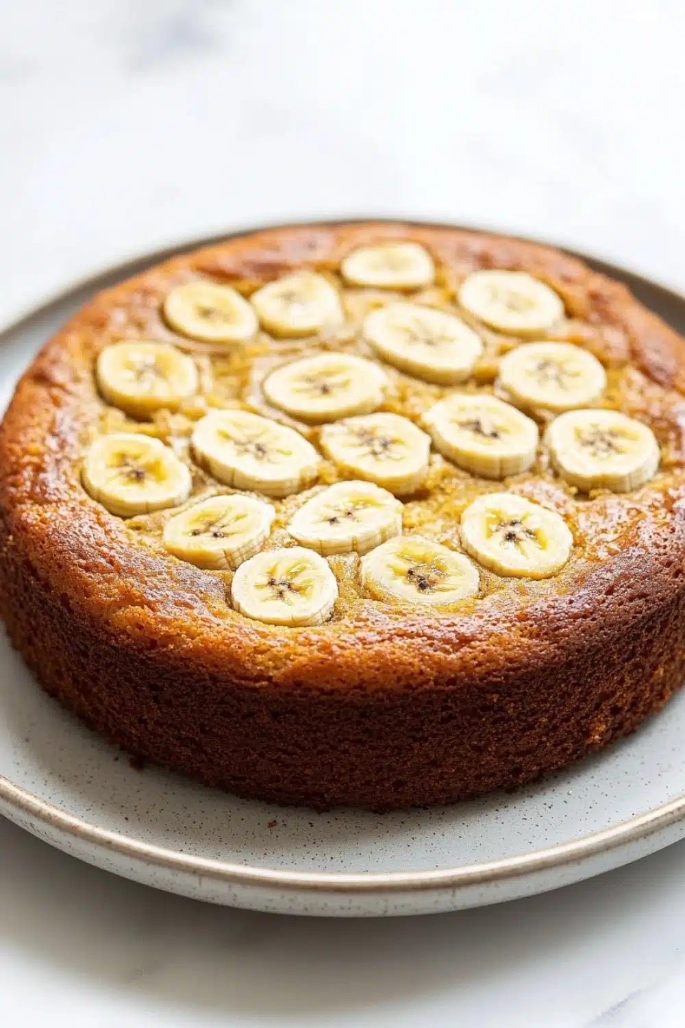 Banana cake - the image is of a round cake on a white plate. the cake appears to be freshly baked and has a golden brown crust. on top of the cake, there are sliced bananas arranged in a circular pattern. the bananas are evenly spaced and appear to be ripe and juicy. the plate is sitting on a marble countertop.
