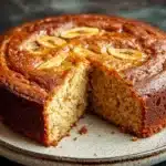 Banana cake - the image shows a round cake on a white plate with a slice taken out of it. the cake appears to be a bundt cake with a golden brown crust and a drizzle of caramel sauce on top. the caramel sauce is drizzled over the top of the cake, creating a swirl pattern. the plate is sitting on a dark grey countertop with a textured surface. the background is blurred, making the cake the focal point of the image.