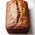 Banana cake loaf photography - the image is a close-up of a freshly baked loaf of bread on a white marble countertop. the bread appears to be golden brown in color and has a slightly crumbly texture. it has a soft, fluffy interior with visible chunks of nuts and seeds scattered throughout. the loaf is slightly tilted to the side, with the top half of the loaf facing towards the right side of the image. the background is blurred, making the bread the focal point of the frame.