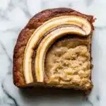 Banana cake slice plating - the image is a close-up of a slice of cake on a white marble surface. the cake appears to be a bundt cake with a golden brown crust and a layer of caramelized bananas on top. the bananas are arranged in a diagonal line, with the top banana being slightly larger than the bottom one. the caramel is drizzled over the top of the cake, creating a smooth and creamy texture. the slice is cut in half, revealing the soft, fluffy interior. the background is blurred, making the cake the focal point of the image.
