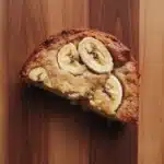 Banana cake slice plating - the image is a close-up of a slice of banana bread on a wooden surface. the bread appears to be freshly baked and has a golden brown crust on top. the slices are arranged in a circular pattern and are evenly spaced along the edges of the bread. the banana slices are cut in half, revealing the soft, fluffy interior. the wooden surface has a rustic, natural look with visible grain and texture. the overall mood of the image is warm and inviting.