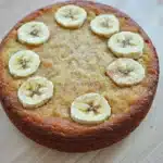 Banana cake toddler - the image is of a round cake with a golden brown crust. on top of the cake, there are several sliced bananas arranged in a circular pattern. the bananas are evenly spaced and appear to be freshly baked. the cake is sitting on a wooden surface, and the background is blurred, making the cake the focal point of the image.