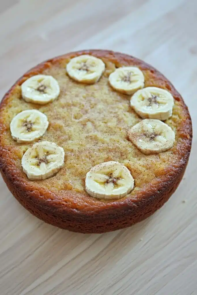 Banana cake toddler - the image is of a round cake with a golden brown crust. on top of the cake, there are several sliced bananas arranged in a circular pattern. the bananas are evenly spaced and appear to be freshly baked. the cake is sitting on a wooden surface, and the background is blurred, making the cake the focal point of the image.