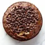 Banana chocolate cake - the image is a close-up of a round chocolate cake on a white marble surface. the cake appears to be freshly baked and has a golden brown color. on top of the cake, there are small chocolate chips scattered all over the surface, creating a textured pattern. the chocolate chips are evenly distributed throughout the cake. the background is blurred, making the cake the focal point of the image.