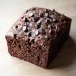 Banana chocolate cake - the image is a close-up of a square-shaped chocolate cake on a wooden surface. the cake appears to be freshly baked and has a rich, dark brown color. it is topped with small, chocolate chips scattered across the top, giving it a shiny appearance. the edges of the cake are slightly curled, indicating that it has been freshly baked. the background is blurred, making the cake the focal point of the image.