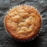 Banana muffin gluten free - the image is a close-up of a freshly baked muffin in a white paper liner. the muffin appears to be golden brown in color and has a crumbly texture. it is sitting on a dark, textured surface with a rough, uneven texture. the edges of the muffin are slightly curled, giving it a textured appearance. the background is blurred, making the muffins the focal point of the image.